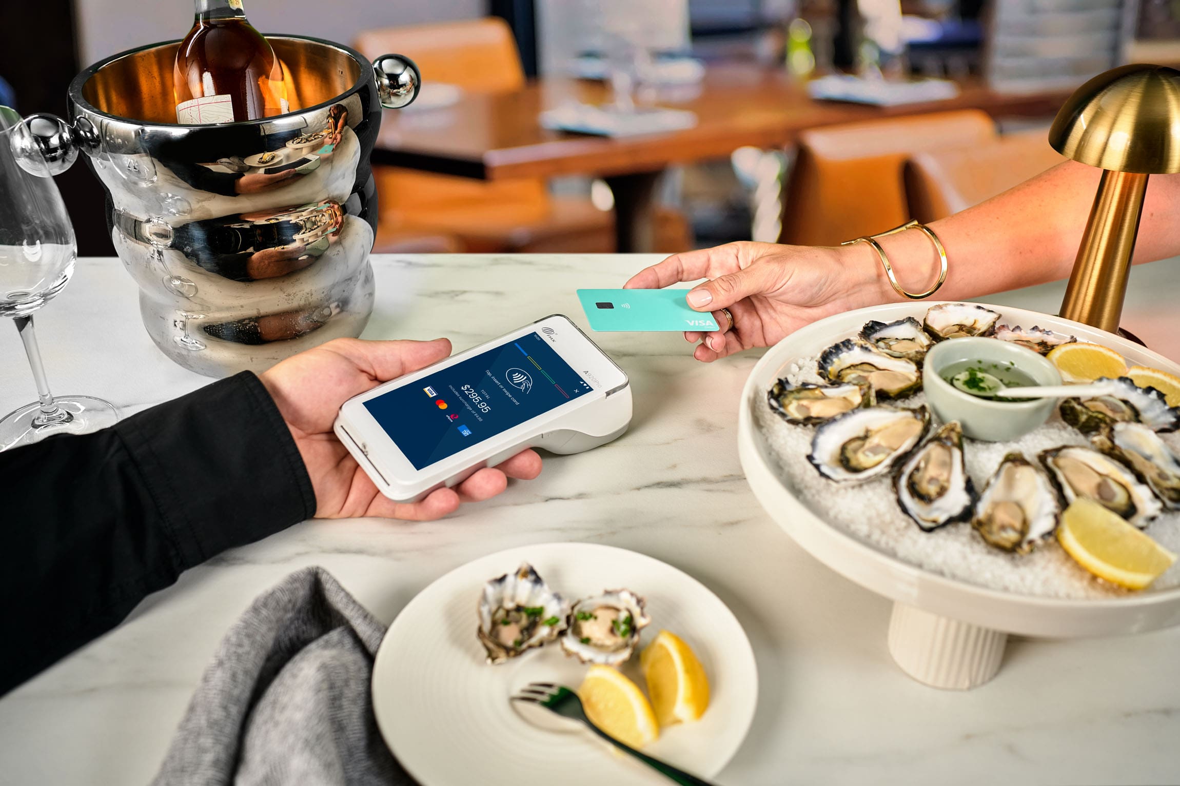 Visa card tapping Android terminal on marble table with plate of oysters and champagne bottle