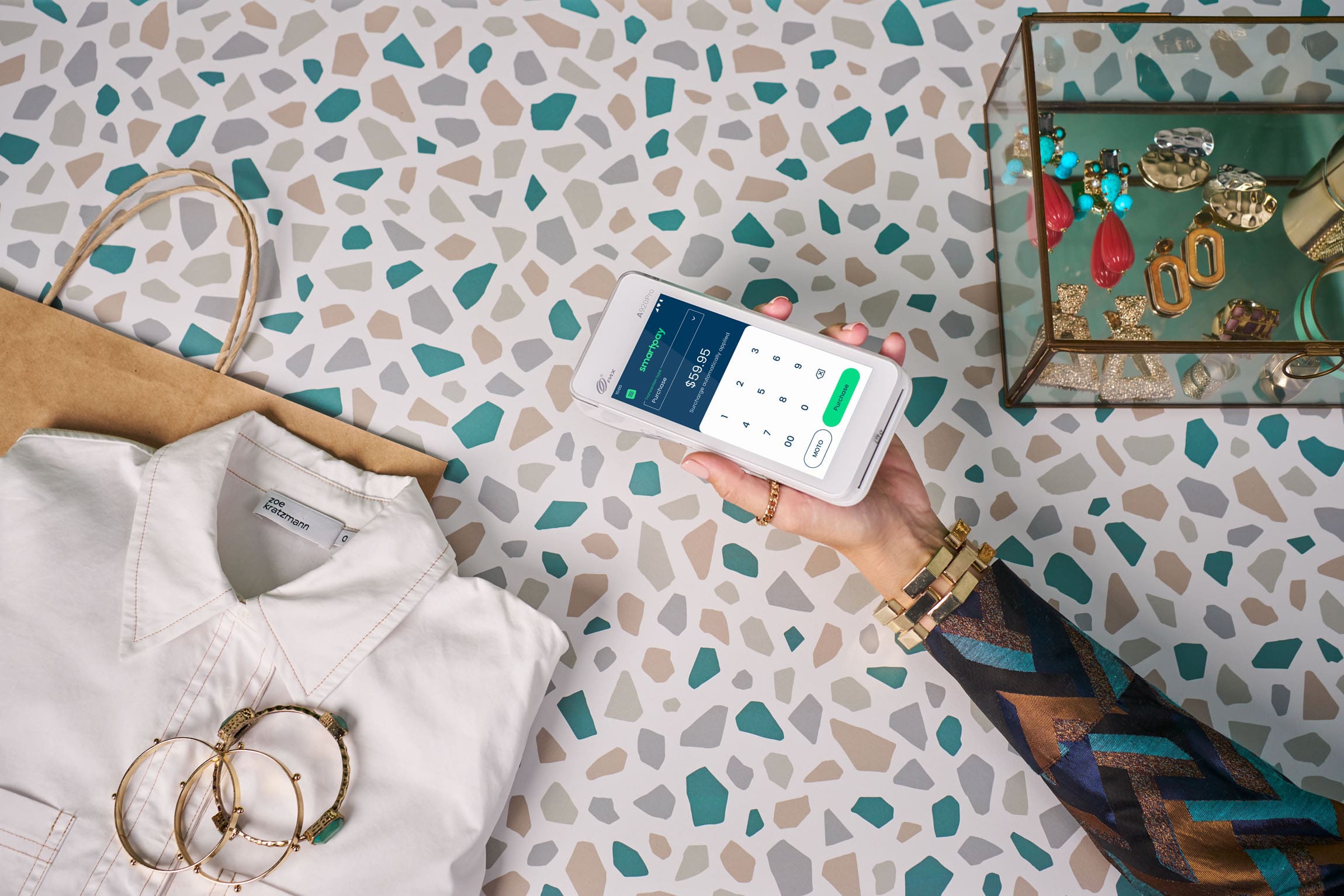 Hand holding a Smartpay terminal over terrazzo counter with clothing and accessories around